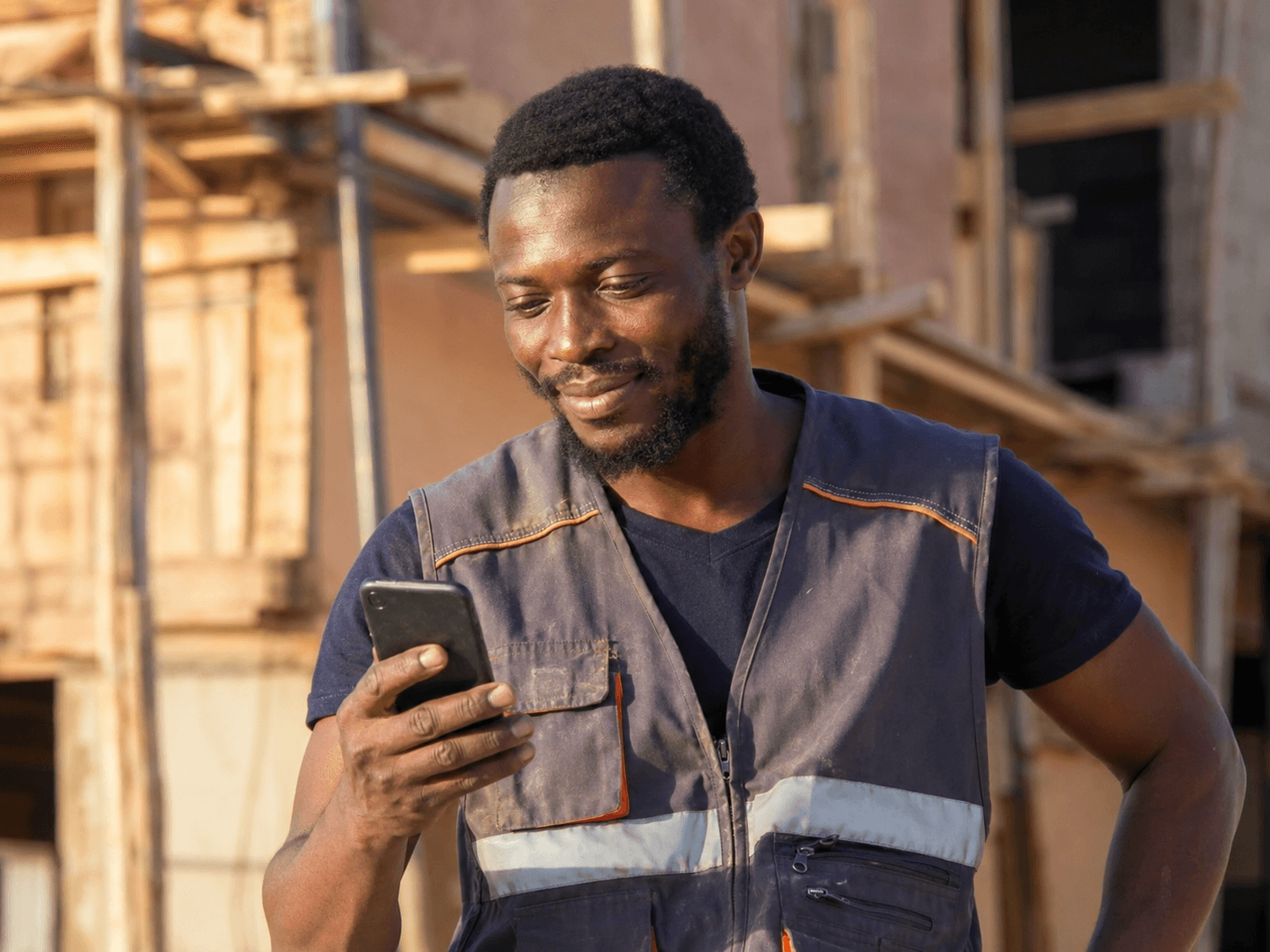 Un artisan en tenue de chantier consulte les demandes reçues sur son téléphone devant un bâtiment en construction.